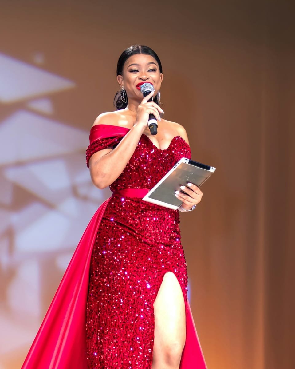 Event host in a red sequined gown smiling as she speaks into a microphone on stage, cue notes in hand